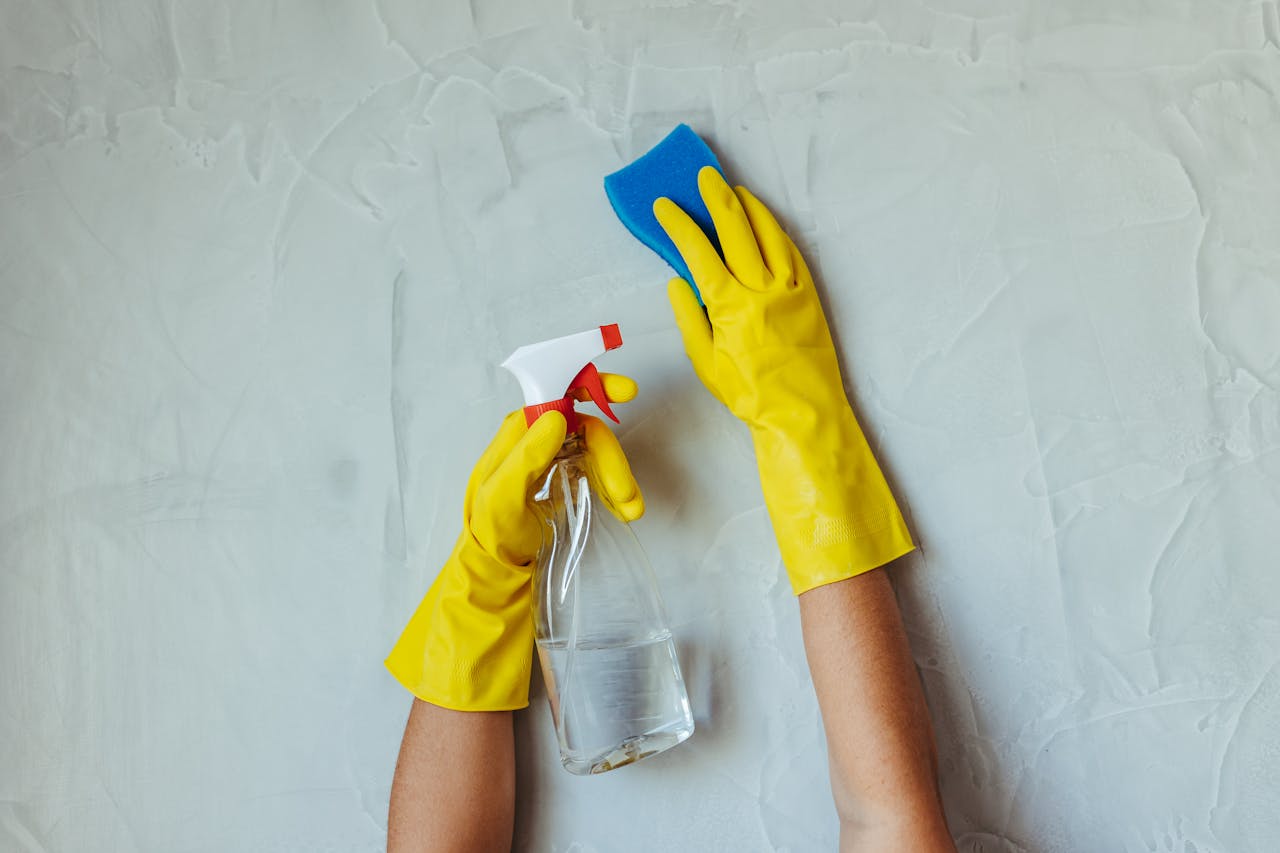 services-03 Close-up of hands in yellow gloves cleaning a wall with a spray bottle and sponge.
