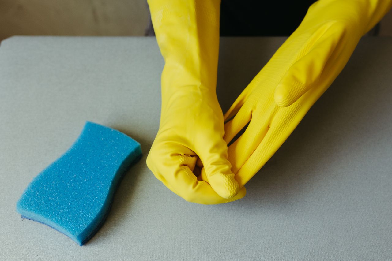 hero-img-02 Close-up of hands wearing yellow gloves next to blue sponge on gray surface.
